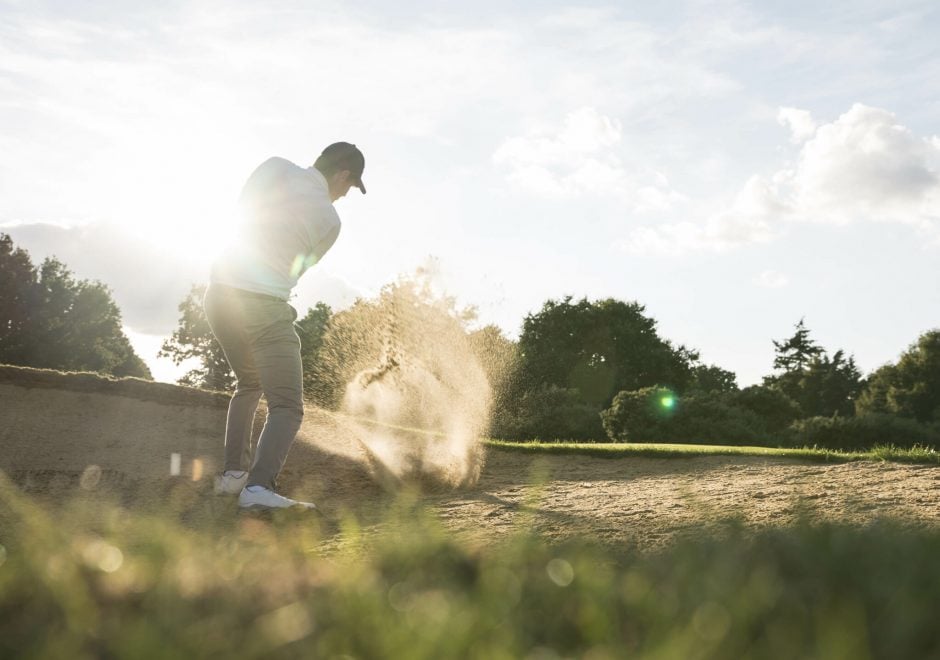 A person swinging at a golf ball on a sand pit under cloudy skies